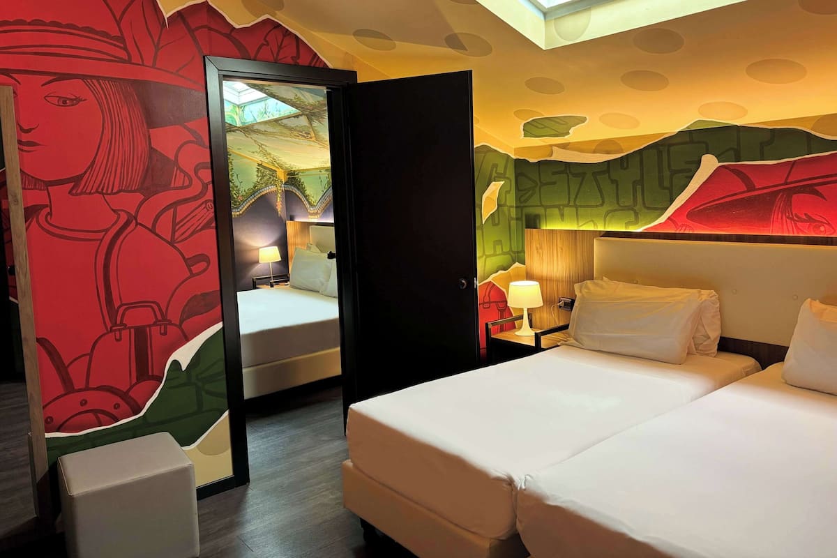 A hotel room with a bed, bedside tables, a mirror, and a wall with a colorful mural.