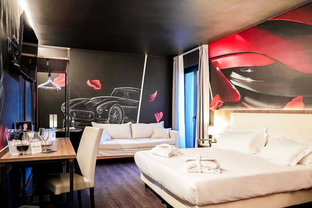 A hotel room with a bed, a sofa, a desk with a chair, and a wall with a car and flower design.