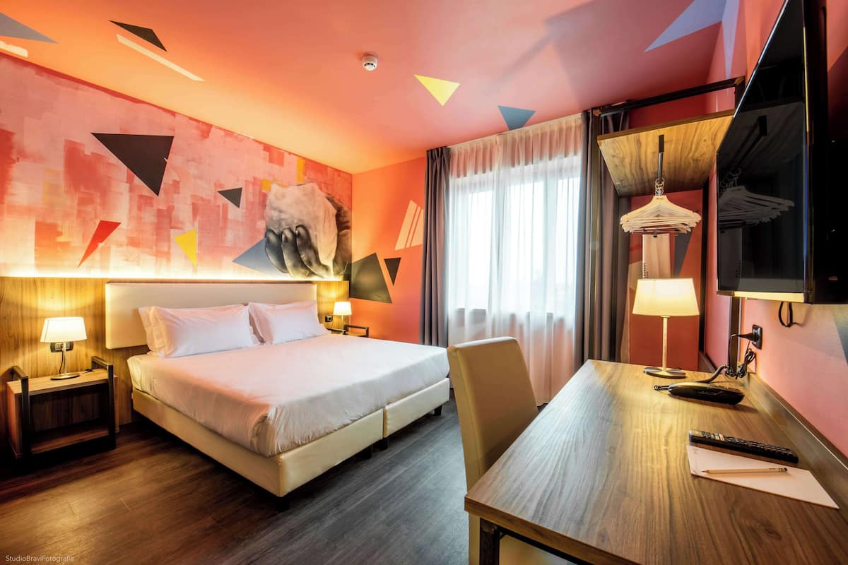 A hotel room with a bed, bedside tables, a desk, a chair, a television, and a window with curtains.