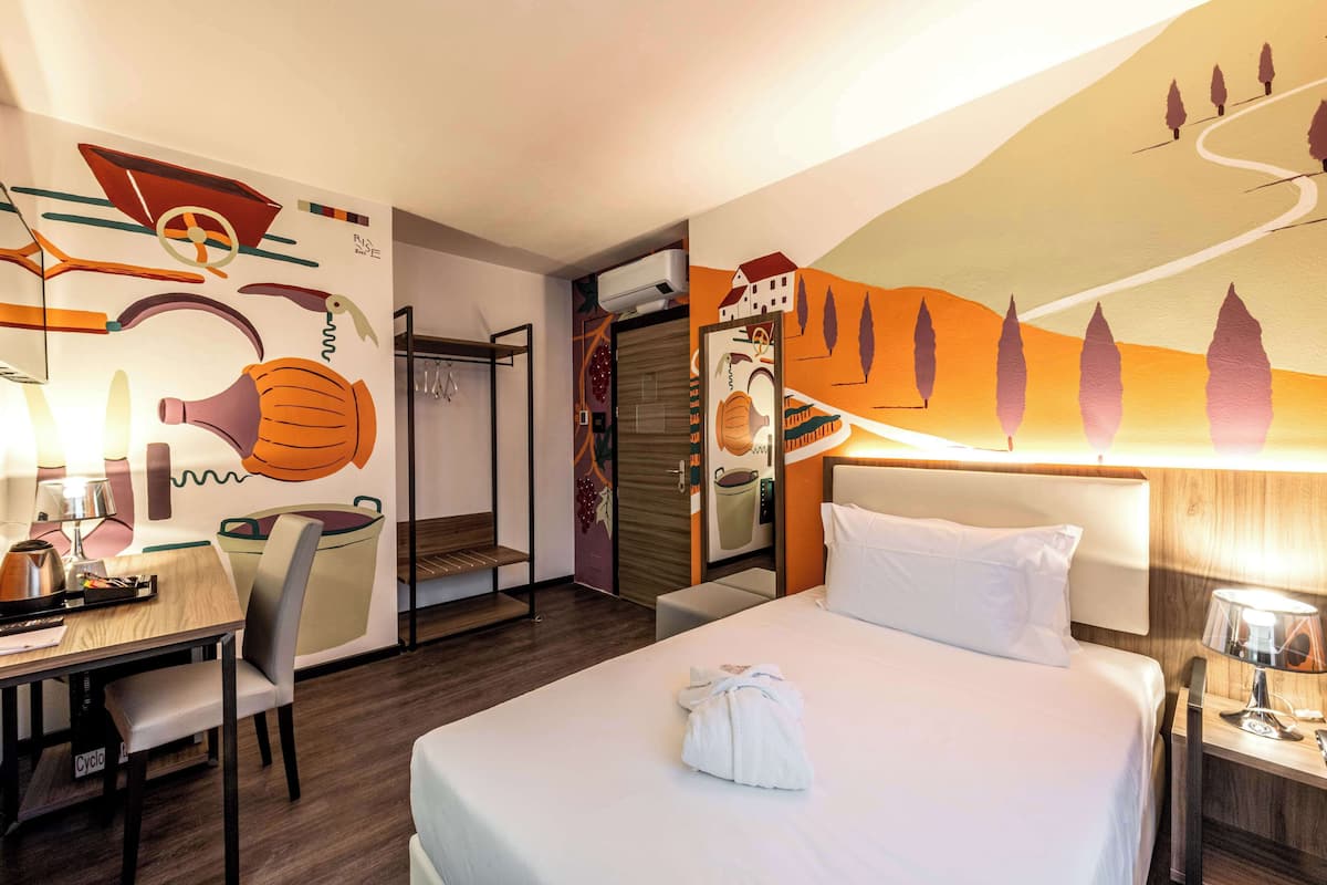 A hotel room with a bed, a desk, and a chair. There is a wall with a mural, a window, and a door.