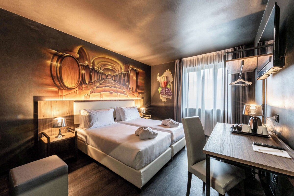 A hotel room with a large bed, two bedside tables, a desk with a chair, a TV, and a wall mural of wine barrels.