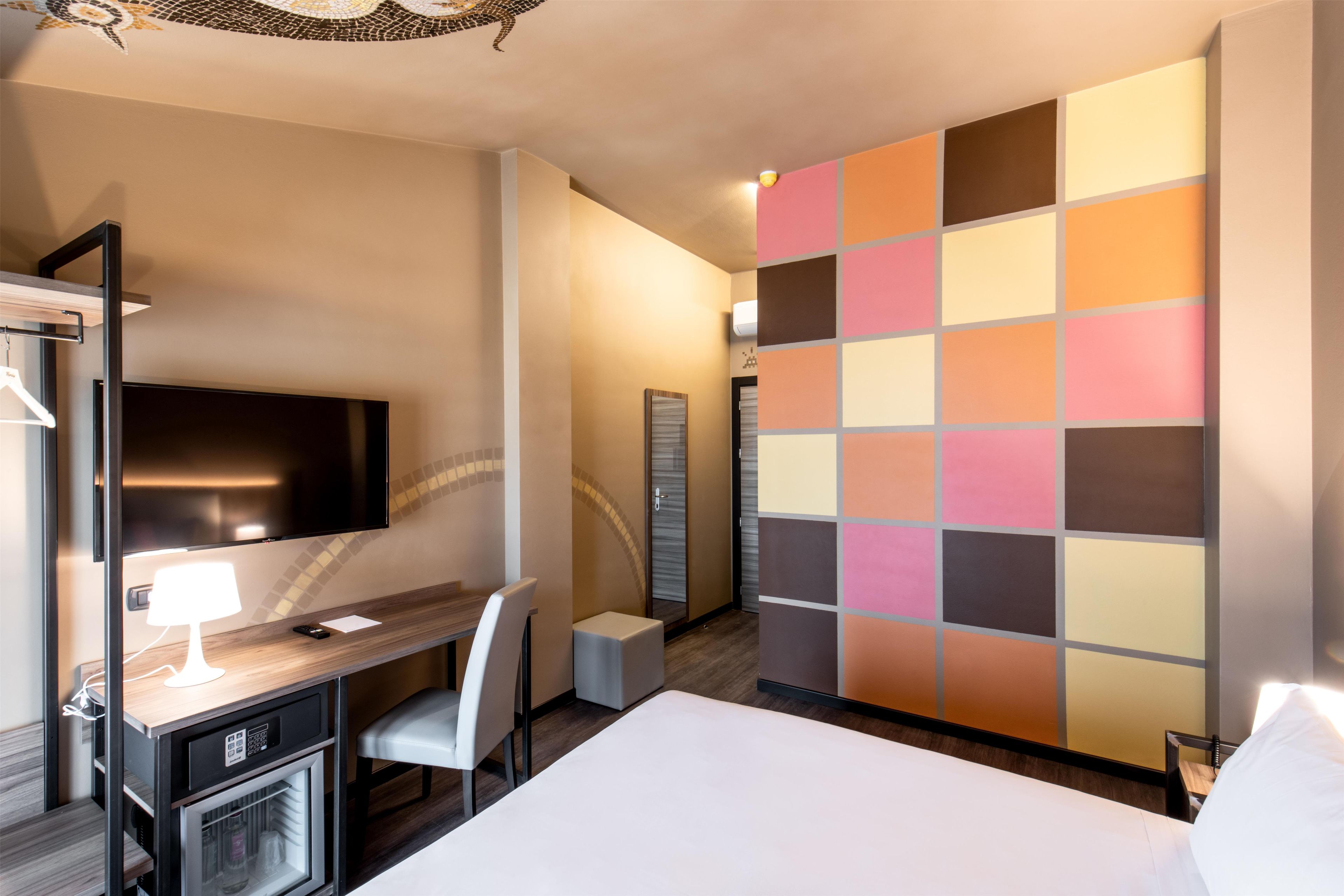 Una habitación de hotel con cama, escritorio con silla, televisor, refrigerador pequeño y un panel decorativo de diseño colorido en la pared.