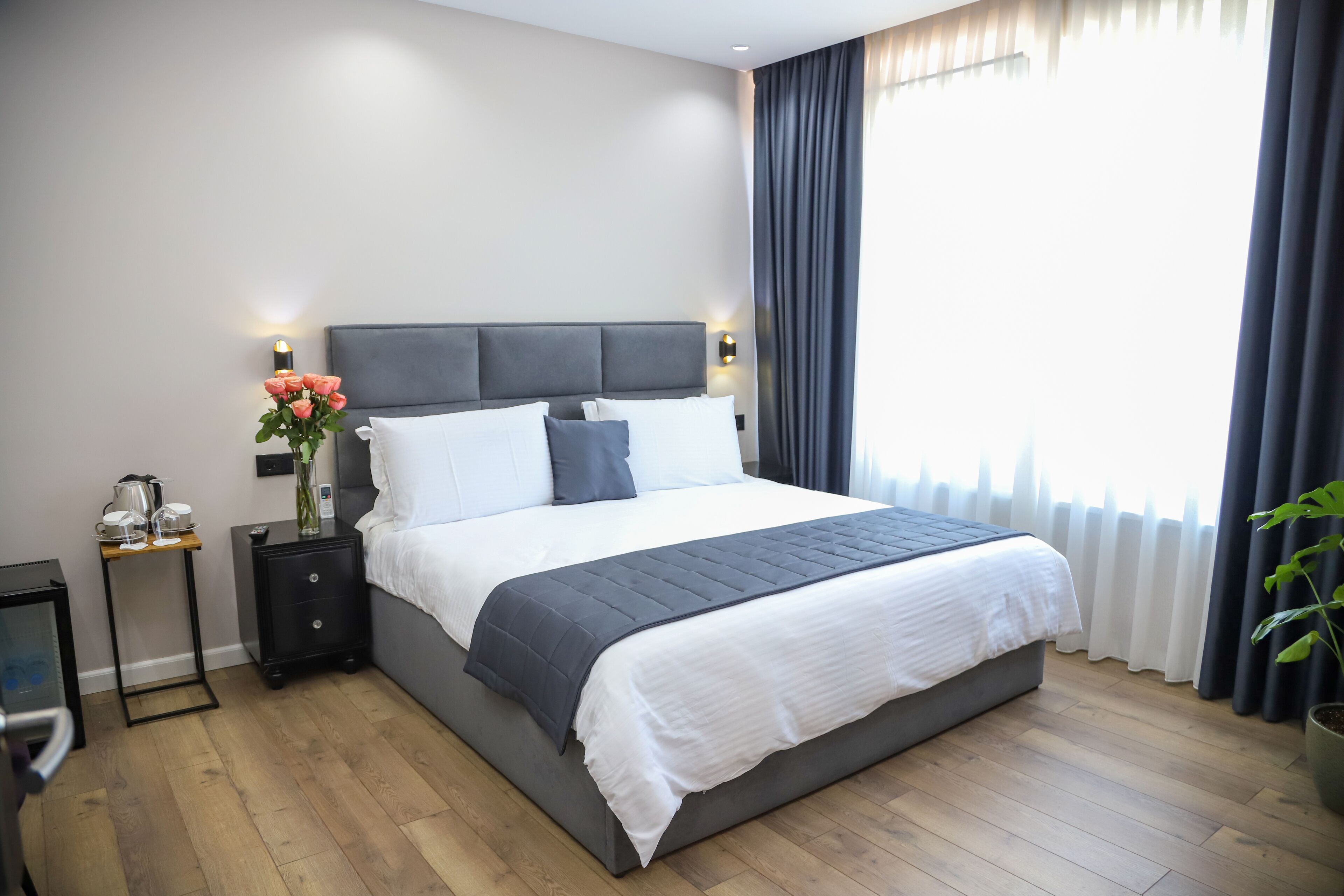 A modern bedroom with a large bed, bedside tables, a nightstand, a vase with flowers, and a window with curtains.