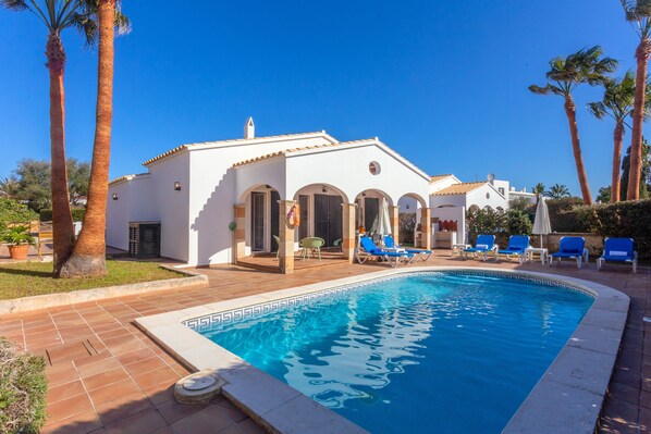 A swimming pool with lounge chairs, a white building with arches, and palm trees.