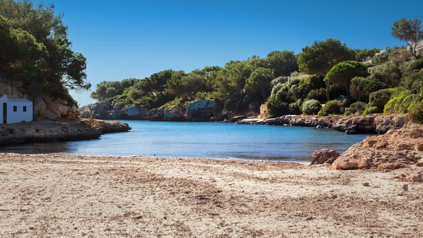 A beach with a white building, clear blue water, and rocky terrain. - 'Villa El Pinar 3' with Pool, Wi-Fi, Garden & Terrace (Ciutadella de Menorca)