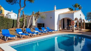 A pool area with blue lounge chairs, a white building with arches, and a clear blue sky. - 'Villa El Pinar 3' with Pool, Wi-Fi, Garden & Terrace (Ciutadella de Menorca)
