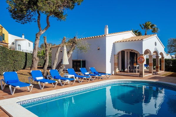 A pool area with blue lounge chairs, a white building with arches, and a clear blue sky.