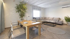 Een moderne woonkamer met een houten eettafel, rieten stoelen, een grijze bank en een grote potplant.