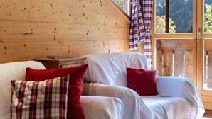 Interior de una cabaña de madera con un sofá y almohadas, una ventana con cortinas y vistas a árboles.