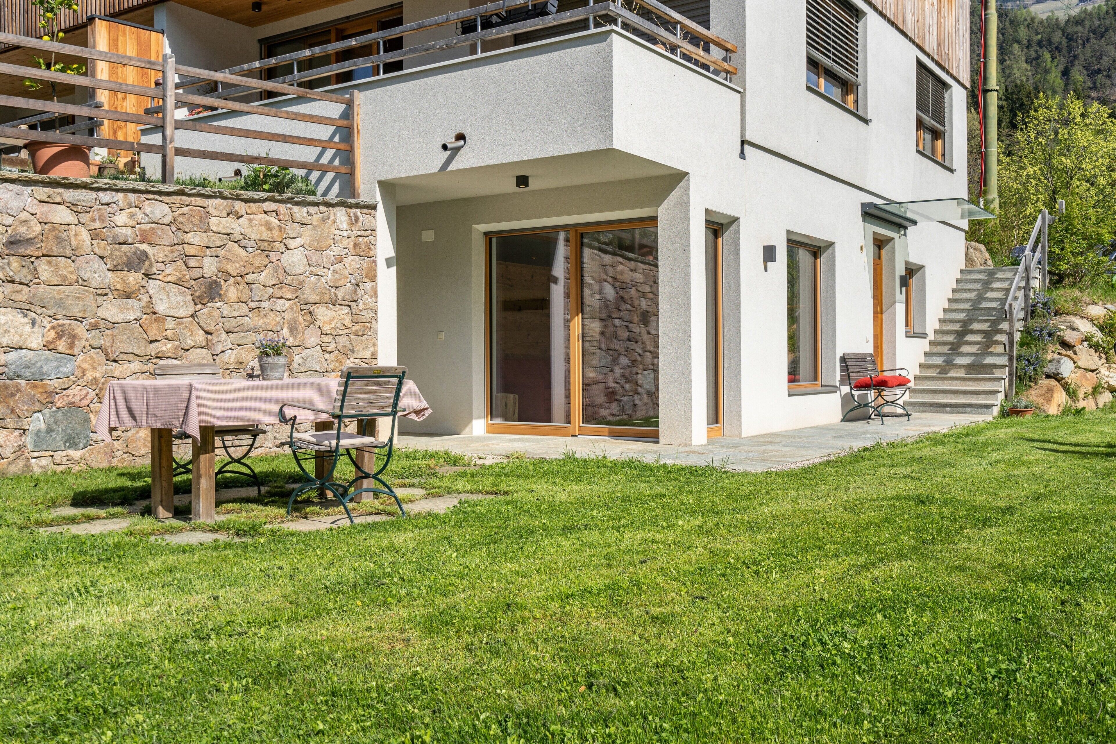 Una casa moderna de dos pisos con un muro de piedra, un porche cubierto con mesa y sillas, y una escalera que conduce al nivel superior.