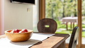 Puisesta pöydästä löytyy kulho omenoita, sen vieressä on tuoli, seinälle on asennettu televisio, ja ikkunasta avautuu näkymä vehreään maisemaan.