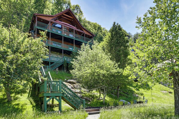 A multi-story log cabin with a balcony, surrounded by lush greenery and a stone staircase.