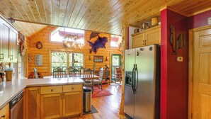 A cabin interior with wooden floors, a kitchen with a refrigerator, wooden cabinets, and a dining area with chairs and a table. - 'River Dream Cabin' on New River w/ Fire Pit (Piney Creek)