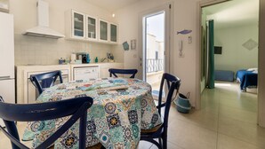 A dining area with a round table, chairs, and a patterned tablecloth. There is a kitchen area with cabinets and a stove. A door leads to a balcony with a view. - Appartamento Playa Iside - Torre... (Torre lapillo)