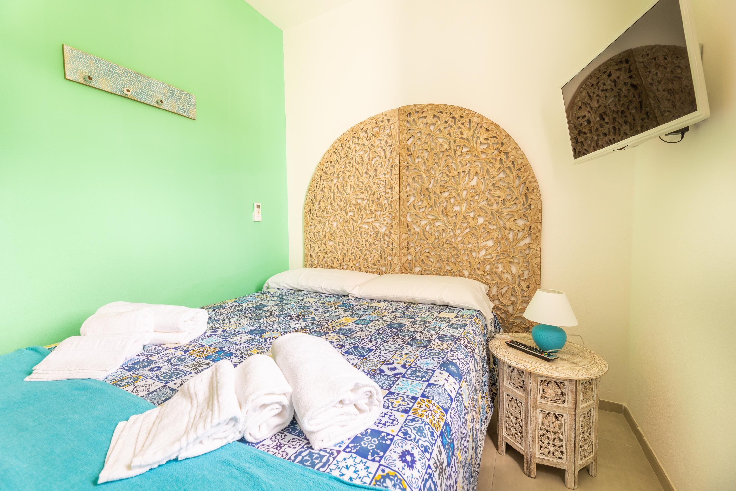 A bed with a patterned bedspread, a headboard with intricate carvings, a bedside table with a lamp, and a wall-mounted TV.