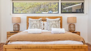 A neatly made bed with a wooden headboard, two bedside tables with lamps, and a large window with a view of trees and a house. - Aiyana.. coastal indulgence at Blueys Beach! (Blueys Beach)