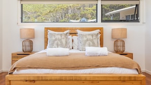 Una cama bien hecha con cabecera de madera, dos mesitas de noche con lámparas y un ventanal con vistas a árboles y una casa.