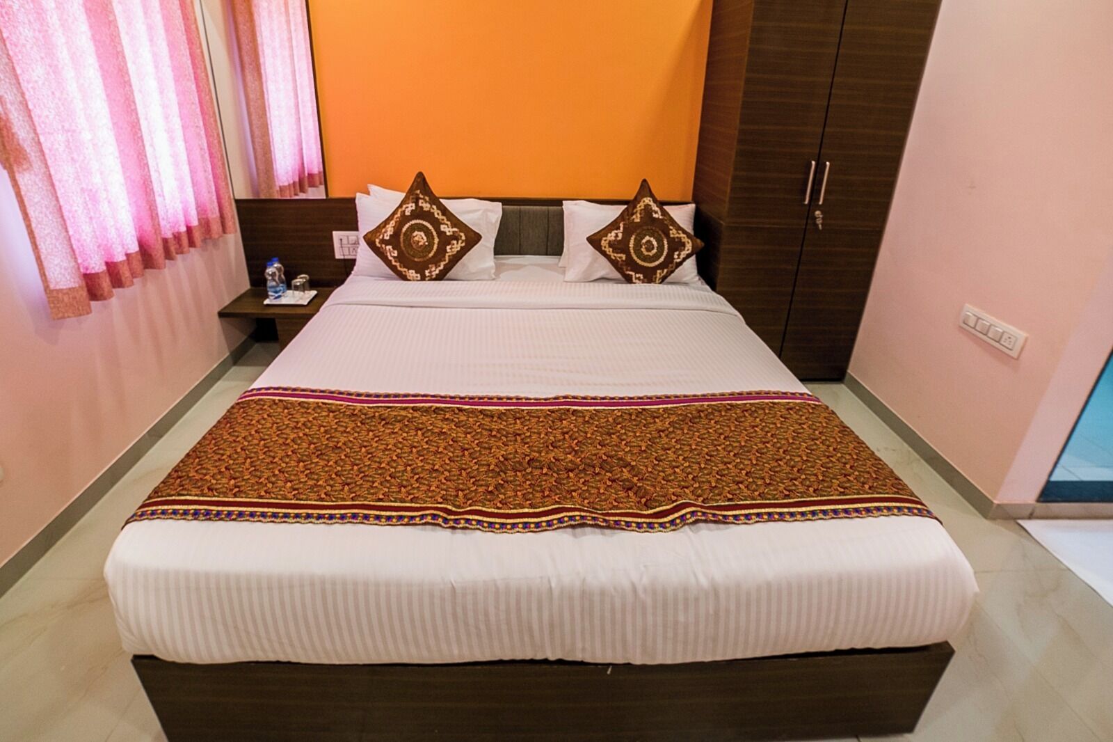 A neatly made bed with a patterned bedspread, two pillows with geometric designs, and a nightstand with a lamp and a bottle.
