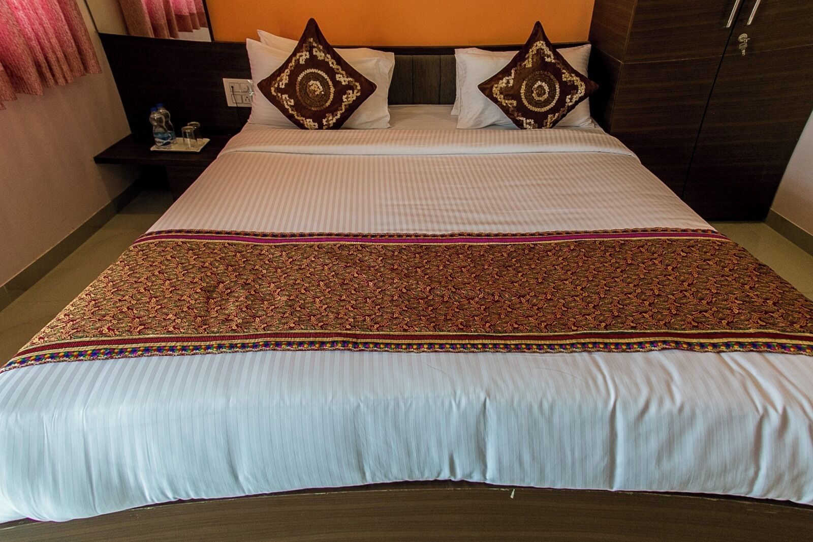 A hotel room with a large bed, two pillows, a nightstand with a water bottle, and a wardrobe.