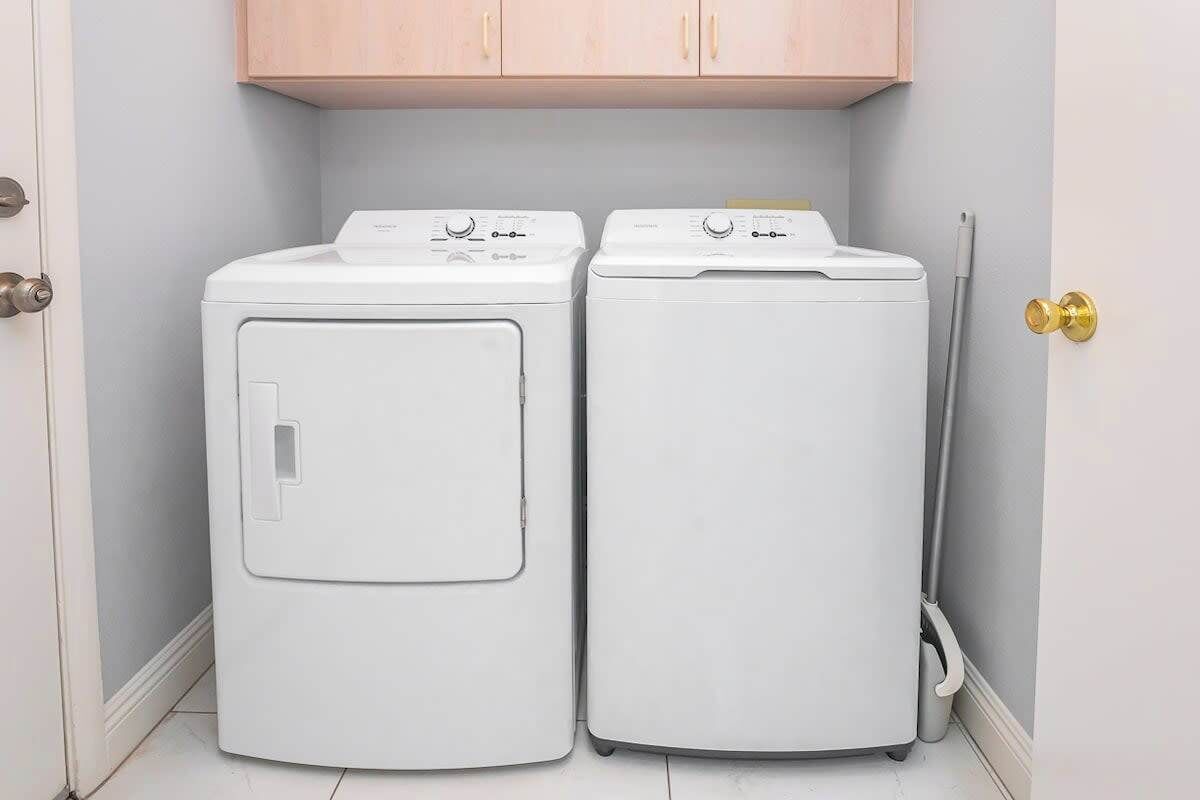Two white front-loading washing machines in a corner with a wooden cabinet above and a mop leaning against the wall.