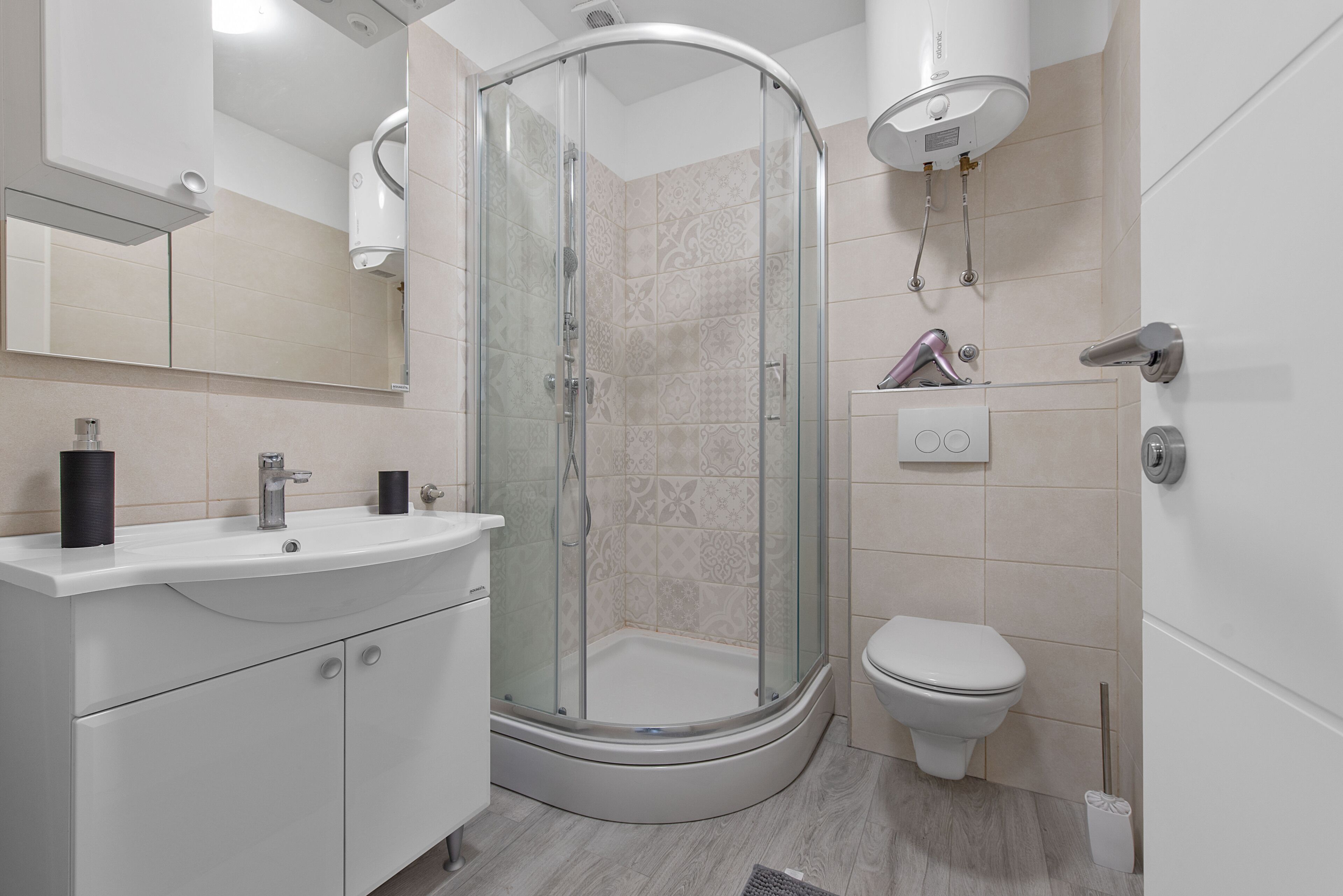 Ein modernes Badezimmer mit einer gläsernen Duschkabine, einem weißen Waschbecken mit zwei schwarzen Seifenspendern und einer Toilette.