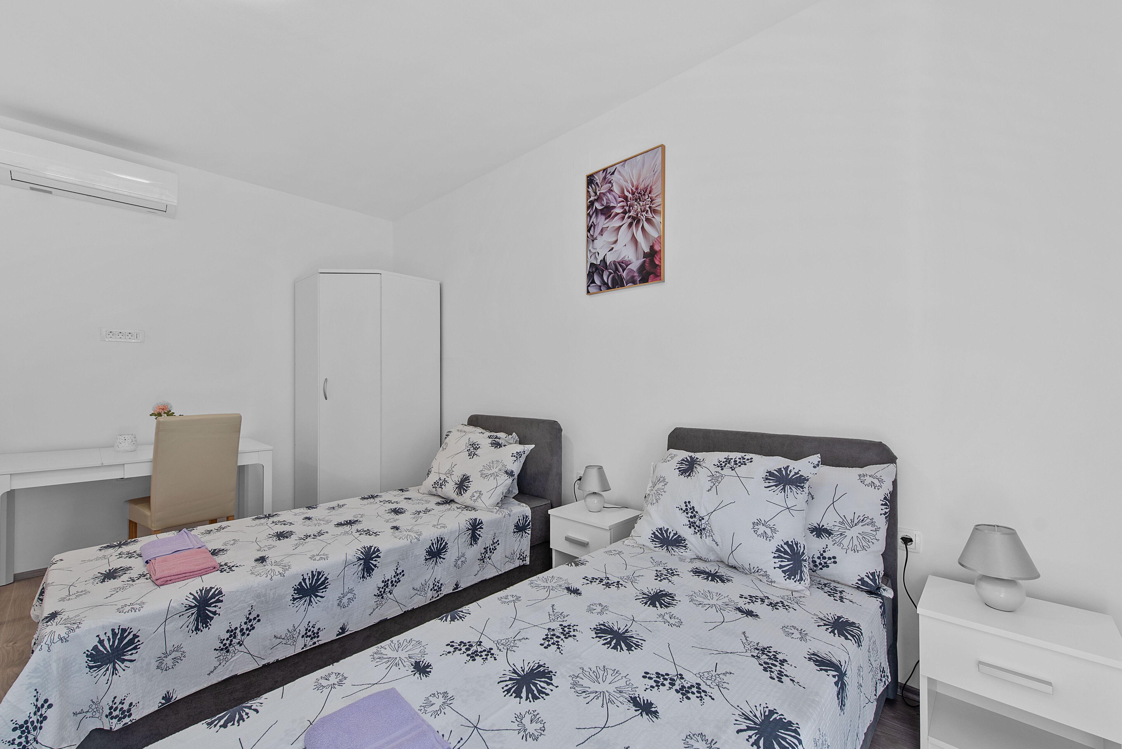 A bedroom with two single beds, a desk, a chair, a wardrobe, and a wall-mounted air conditioner.
