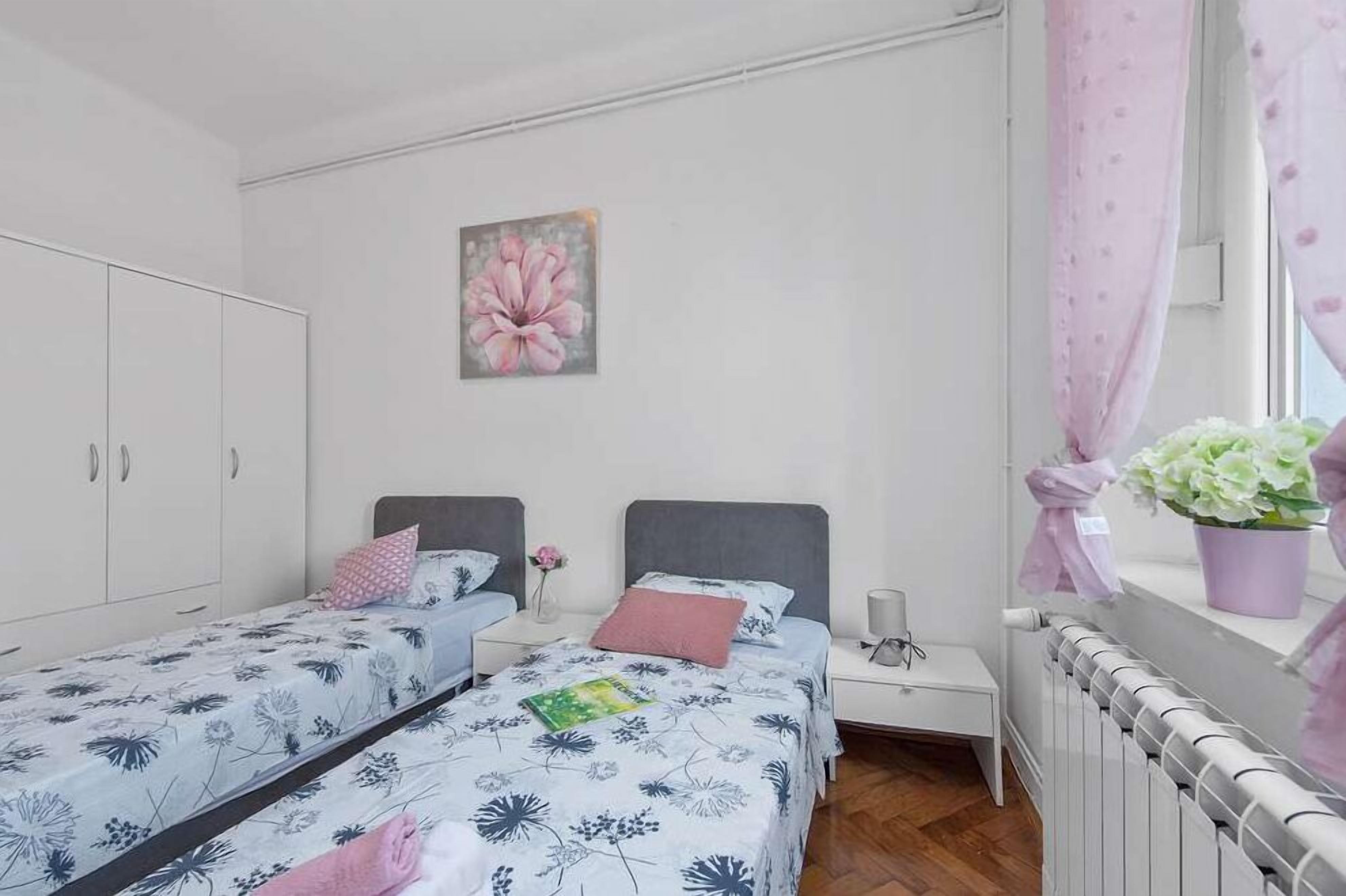 A bedroom with two beds, a white wardrobe, a nightstand with a lamp, and a window with curtains.