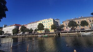 A river with buildings along the riverbank, including a hotel with a red roof and a modern building with a blue sign. - La Guardia apartments (Rijeka)