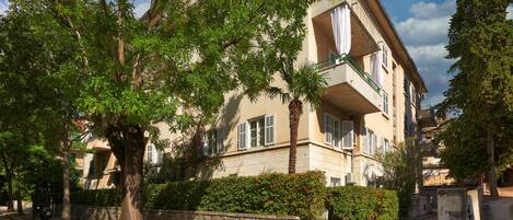 A multi-story residential building with balconies, surrounded by trees and a stone wall.