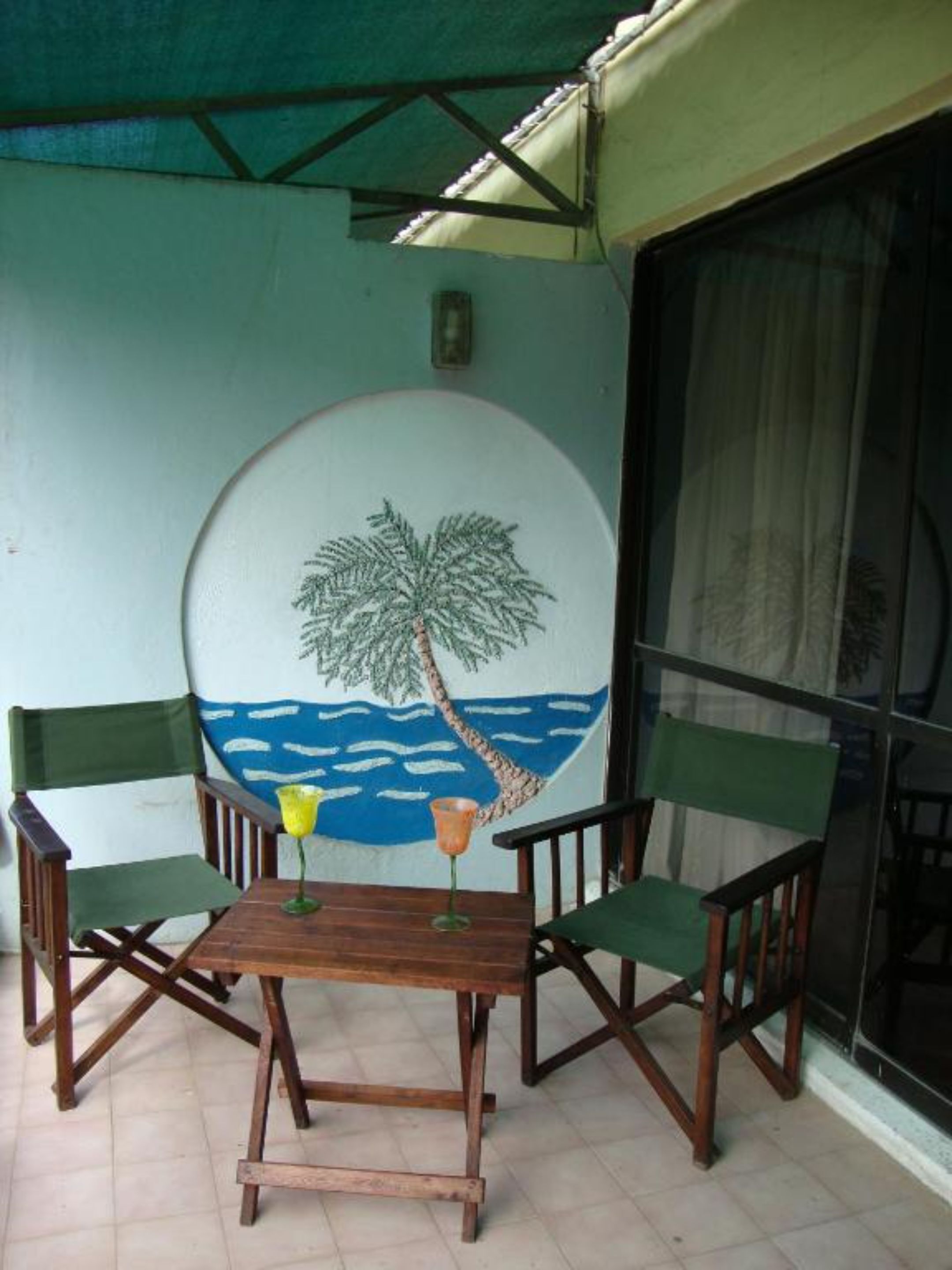 Un petit espace extérieur aménagé avec une table en bois et quatre chaises, une fresque représentant un palmier et l’océan sur le mur, et une fenêtre avec un rideau.