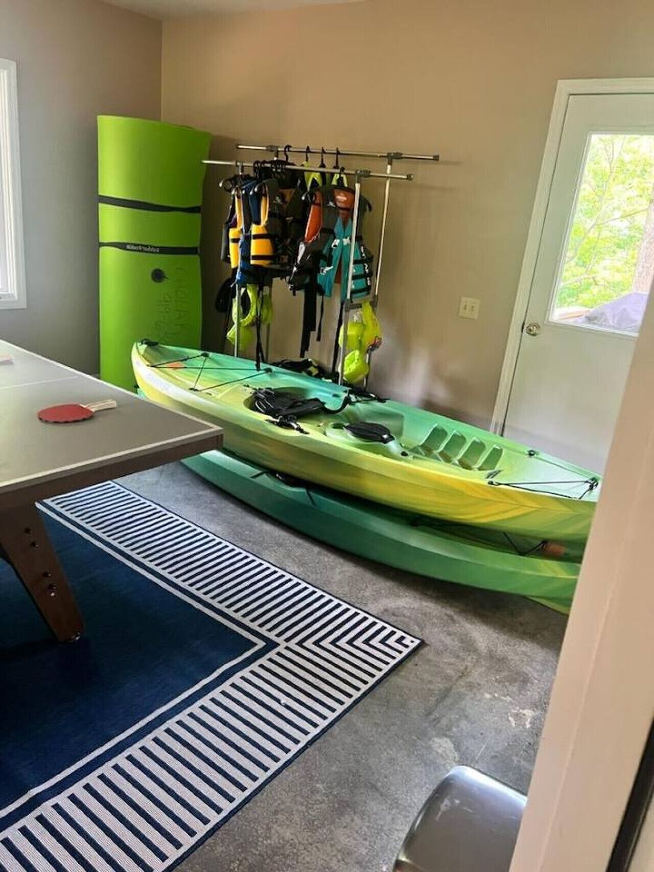 A kayak in a room with a table and a chair, a rack with life jackets, and a door with a view.