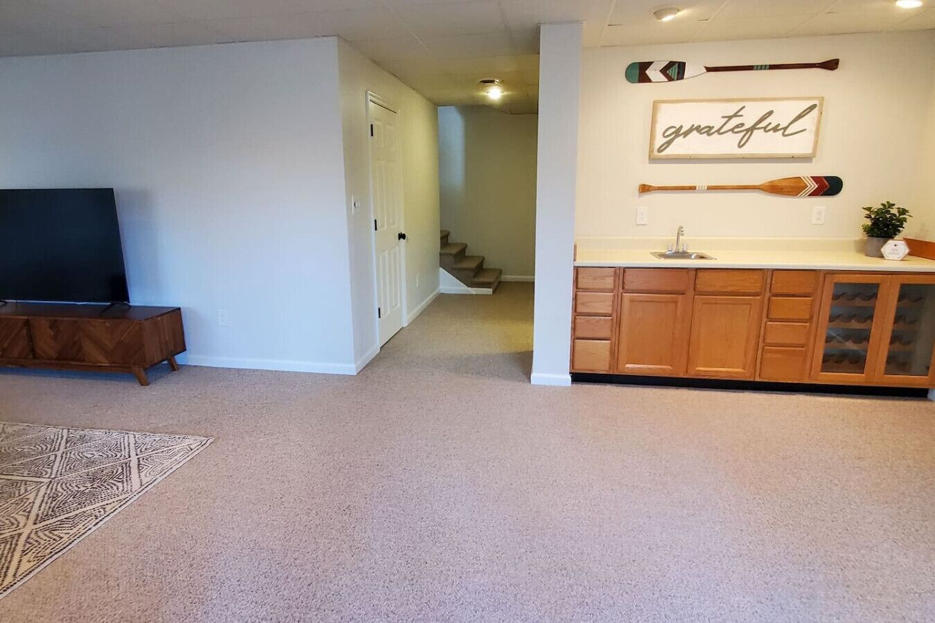 A living room with a TV, a staircase, and a kitchen area with wooden cabinets and a 'grateful' sign.
