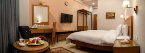 A hotel room with a bed, a desk with a chair, a television, a mirror, and a dining table with fruits and a kettle.
