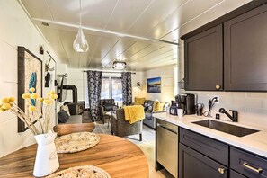 Eine moderne Küche mit dunklen Schränken, einer weißen Arbeitsplatte und einem Spülbecken. Es gibt einen Essbereich mit einem Holztisch und Stühlen, sowie ein Wohnzimmer im Hintergrund.