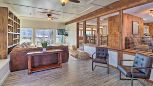 A spacious living area with a large sofa, a coffee table, and two chairs. There are ceiling fans, a fireplace, and a view of the outside through large windows.