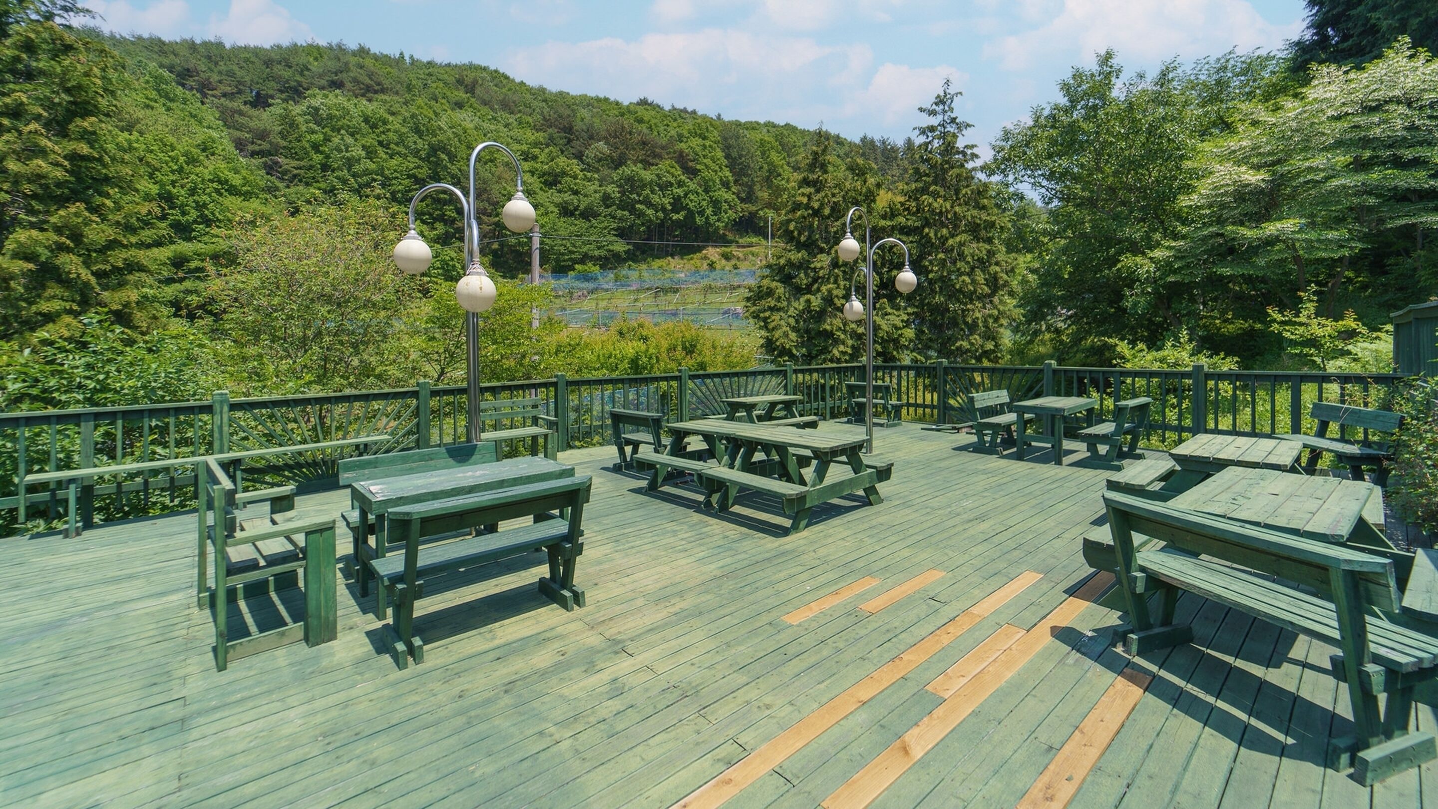 A wooden deck with picnic tables and benches, surrounded by a green railing and trees.