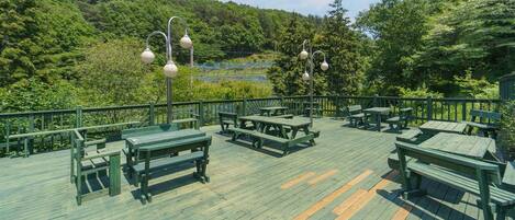 A wooden deck with picnic tables and benches, surrounded by a green railing and trees.