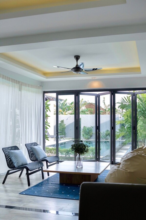 A modern living room with a large sliding glass door leading to a pool area, a ceiling fan, and a coffee table with a vase. - Blu Boat Luxury Pool Villas (Choeng Thale)