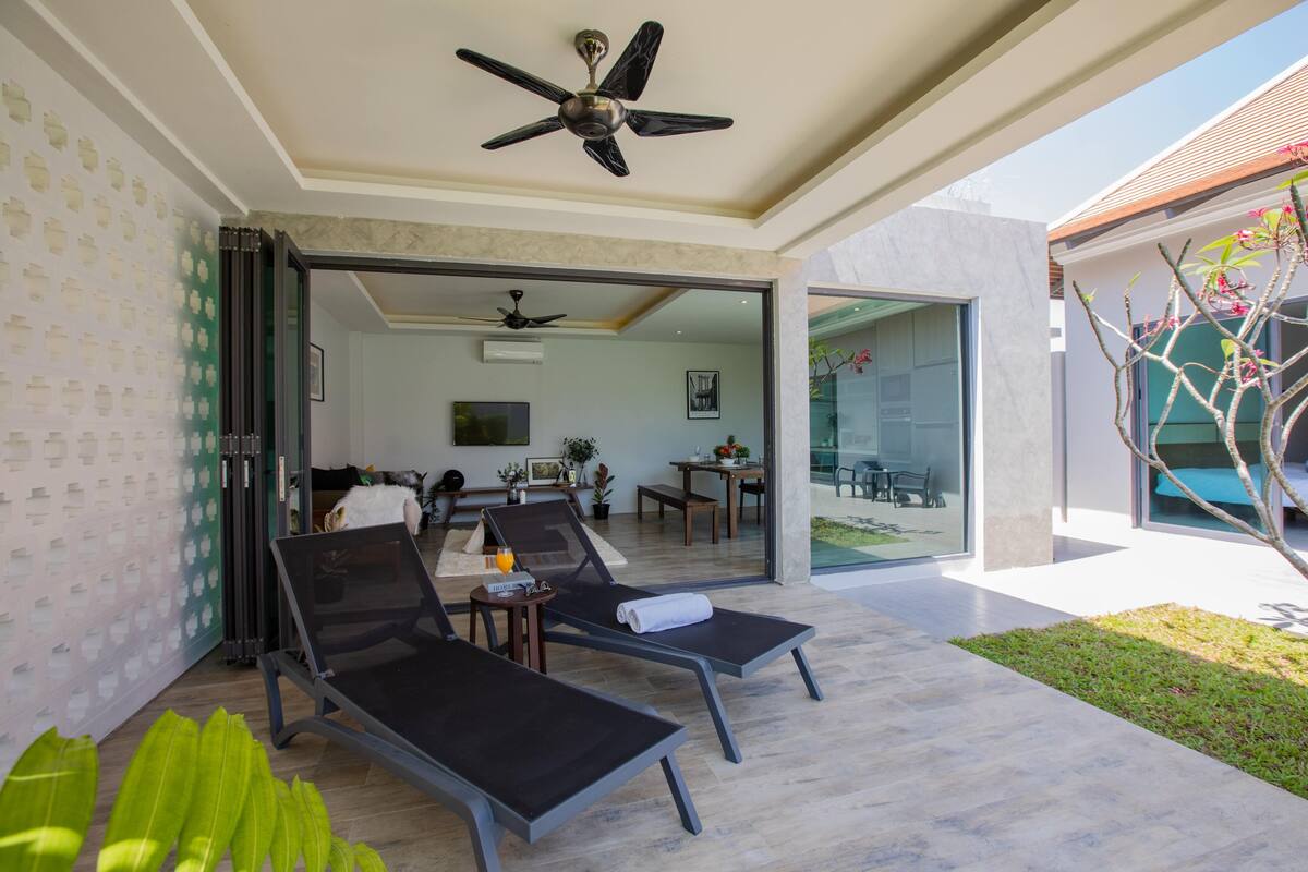Un espacio lounge exterior moderno con dos tumbonas, un ventilador de techo y un ventanal de vidrio que da a un comedor interior.