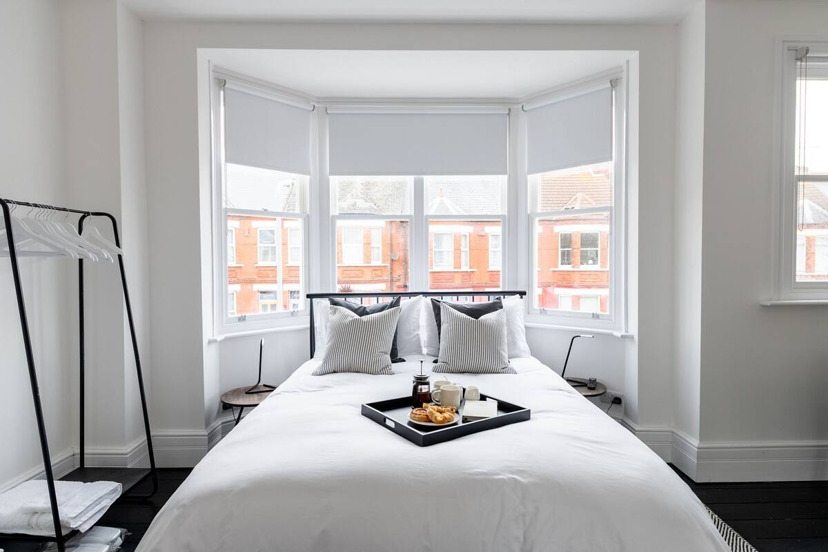 Cama bem arrumada com uma bandeja de itens de café da manhã, um varal de metal preto e vista para edifícios de tijolos vermelhos através de grandes janelas.