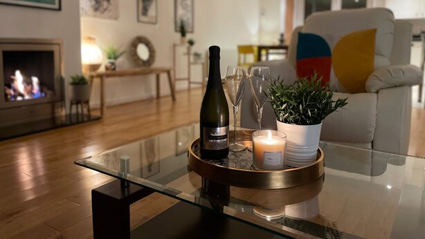 A modern living room with a glass coffee table holding a bottle, glasses, and a candle. A fireplace is visible in the background, and there are framed artworks on the wall. - Barn End, 9 Stad Clynnog (Llanfairpwllgwyngyll)