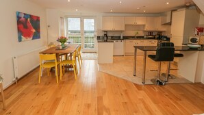 A modern kitchen with wooden flooring, a dining area with yellow chairs, and a view through a glass door to an outdoor balcony. - Barn End, 9 Stad Clynnog (Llanfairpwllgwyngyll)