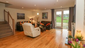 A spacious living room with a staircase, a large window with curtains, and a view of the outdoors. - Barn End, 9 Stad Clynnog (Llanfairpwllgwyngyll)
