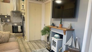 Een gezellige woonkamer met een bank, een open haard met decoratieve objecten, een televisie en een keuken met apparatuur.