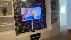 Sala de estar com televisão de tela plana na parede, papel de parede floral, um móvel branco embaixo e um sofá bege.