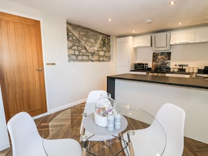 A modern kitchen with white cabinets, a black countertop, and a stone backsplash. There is a dining area with a glass table and white chairs. - The Chalet (Tyn-y-Gongl)