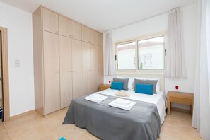A bedroom with a large wardrobe, a bed with grey bedding, blue pillows, and a window with curtains.