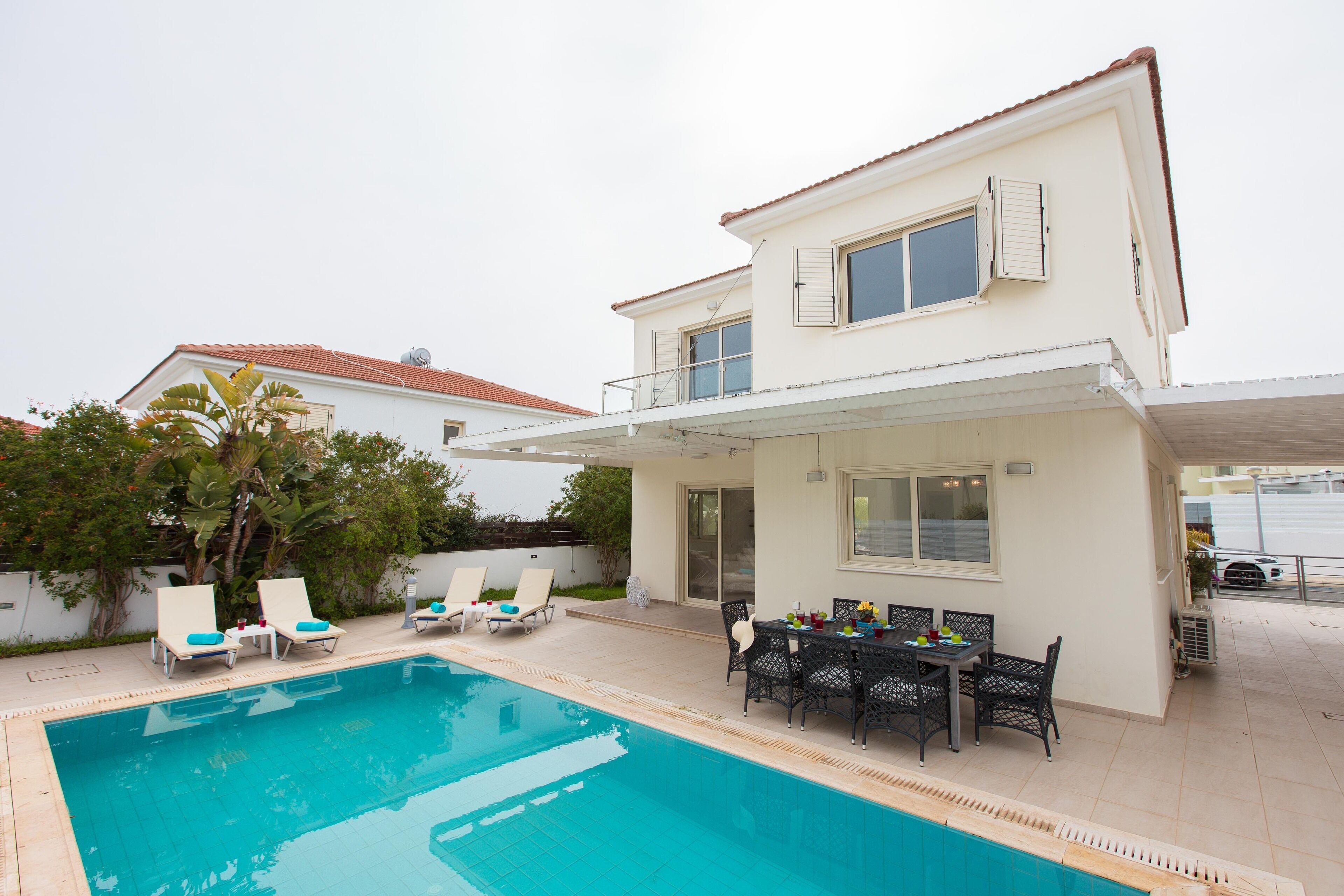 Una casa de dos pisos con piscina, mobiliario exterior y un patio cubierto.
