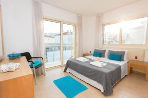 A modern hotel room with a bed, a desk, a chair, and a view of the city through large windows.