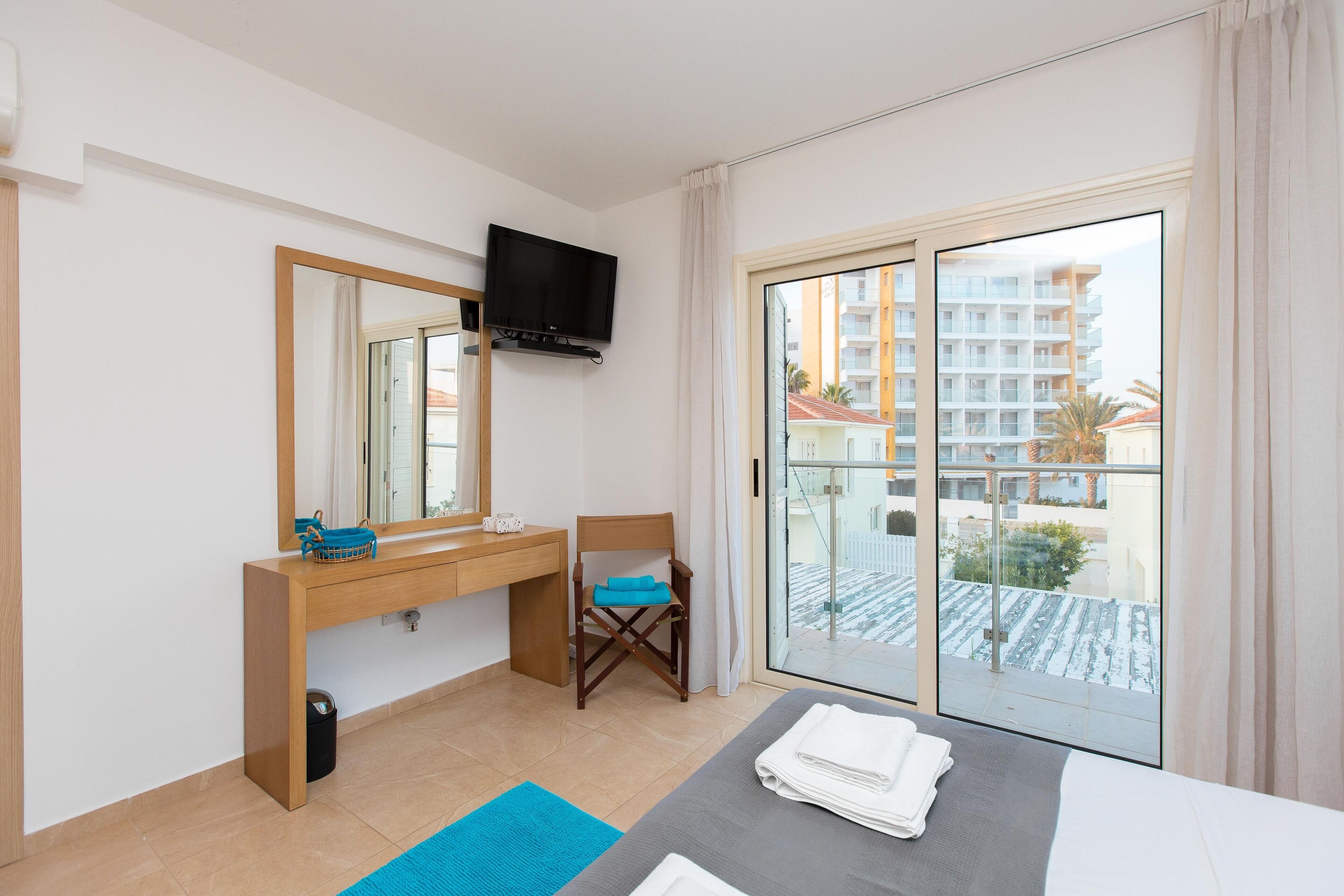 Et hotelværelse med seng, skrivebord med spejl, stol, fjernsyn ophængt på væggen og en balkon med udsigt til bygninger.